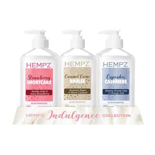 Three HEMPZ pump bottles from the Indulgence Collection arranged on a display. The bottles are labeled Strawberry Shortcake, Caramel Crème Brûlée, and Cupcakes Cashmere, with pink, beige, and blue labels respectively.